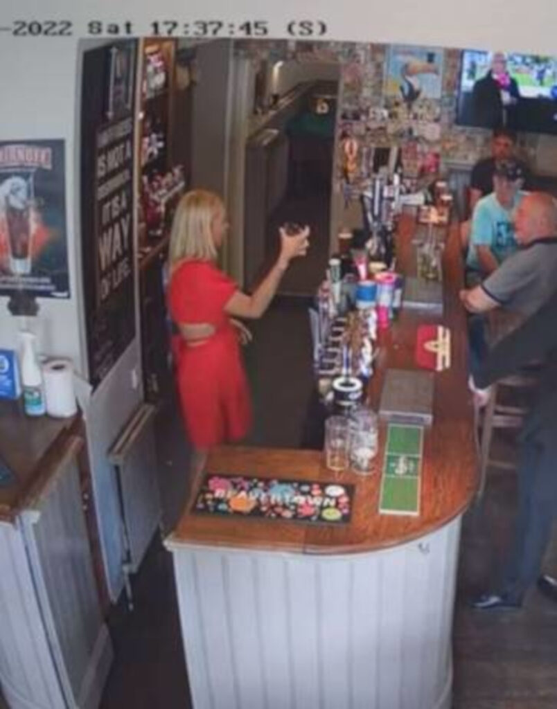 A woman stood behind a bar holding half of a pint glass.