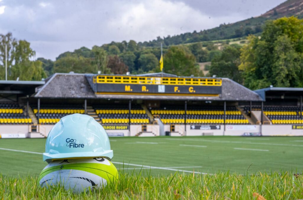 A rugby ball sitting on a field with a Gofibre helmet on top of it - Melrose Rugby Club can be seen in the background. 