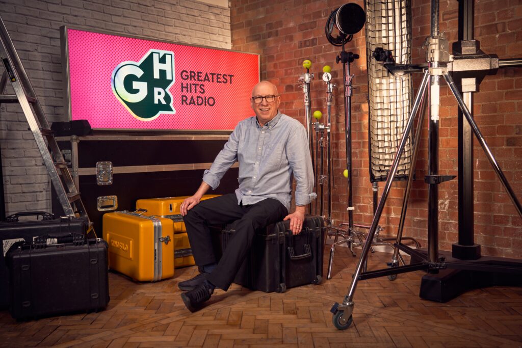 Radio presenter Ken Bruce poses in front of the Greatest Hits Radio logo.