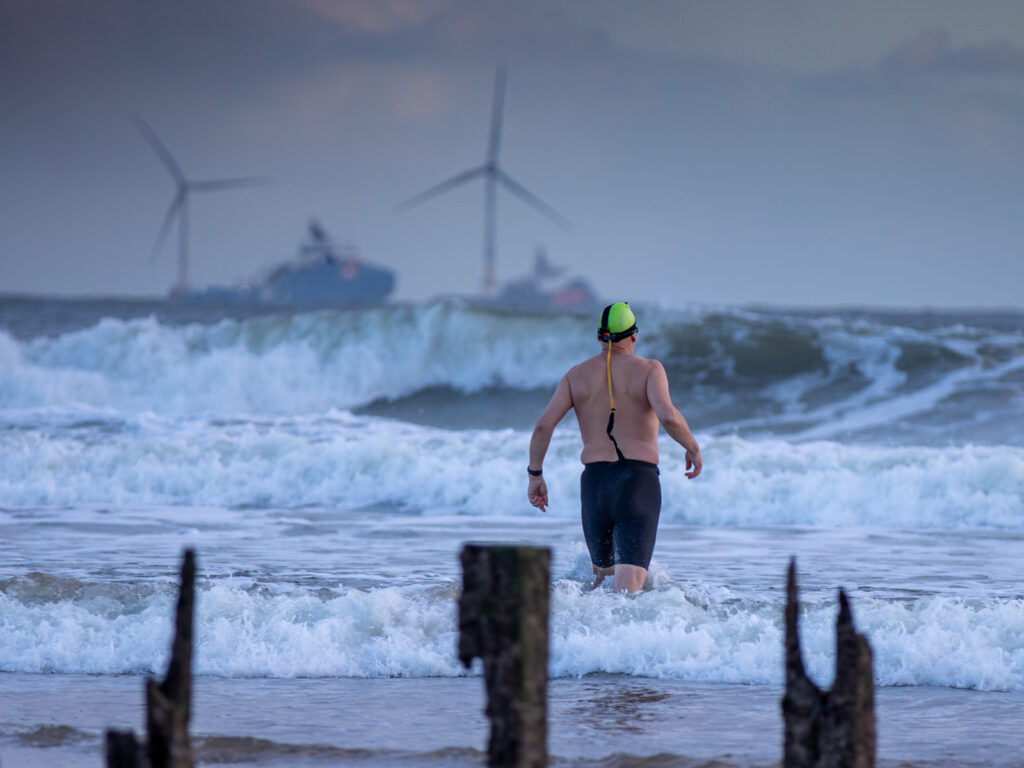 A man walking into the sea with an offshore wind farm in the background.