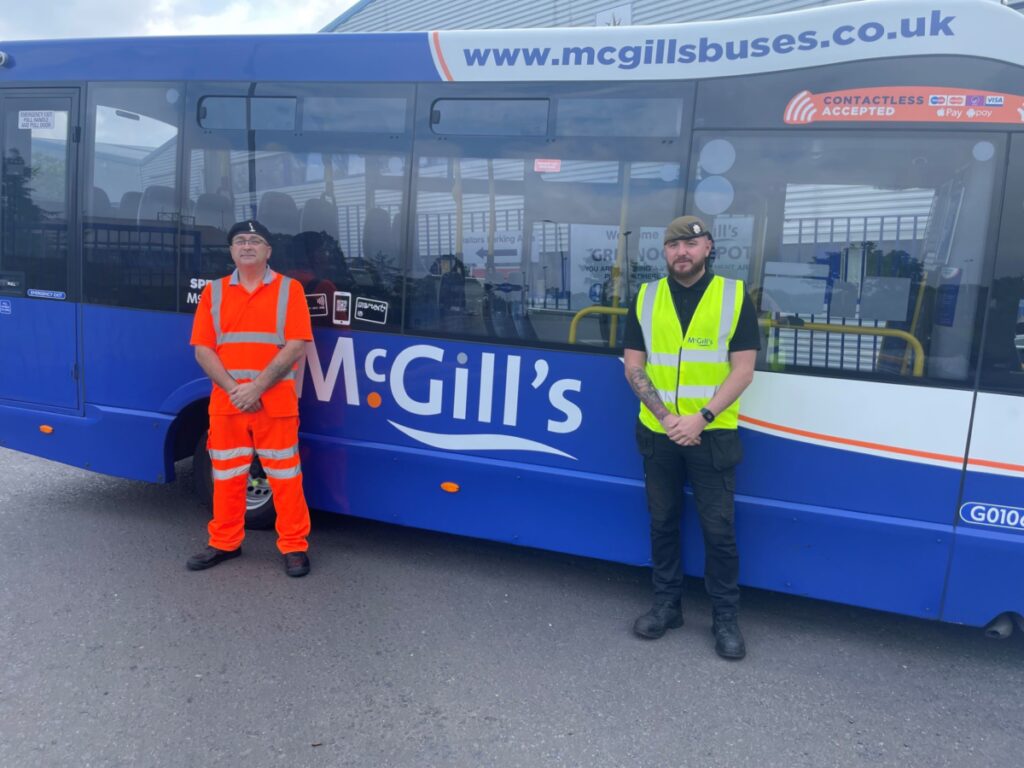Two veterans in hi vis jackets, stood in front of a blue McGills bus