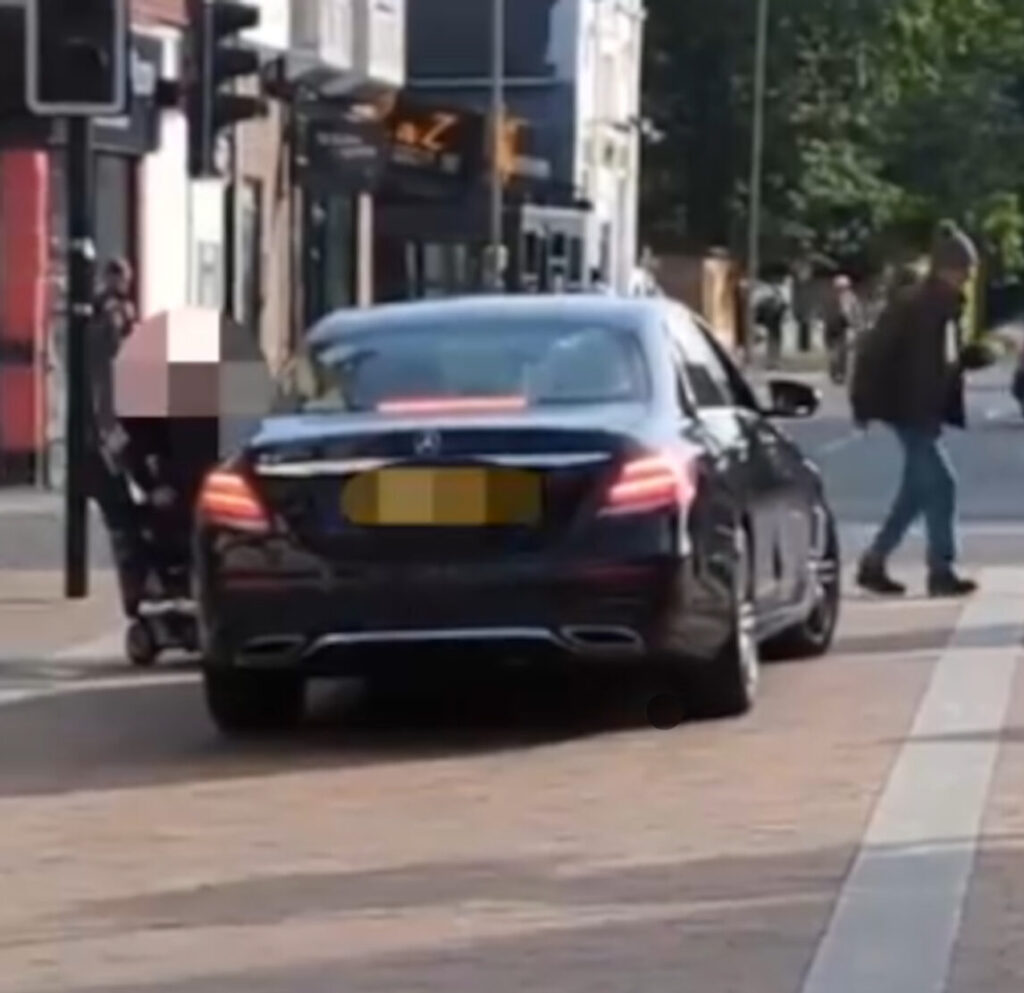 A Mercedes car stopped at a red traffic light whilst a woman on a mobility scooter leans in