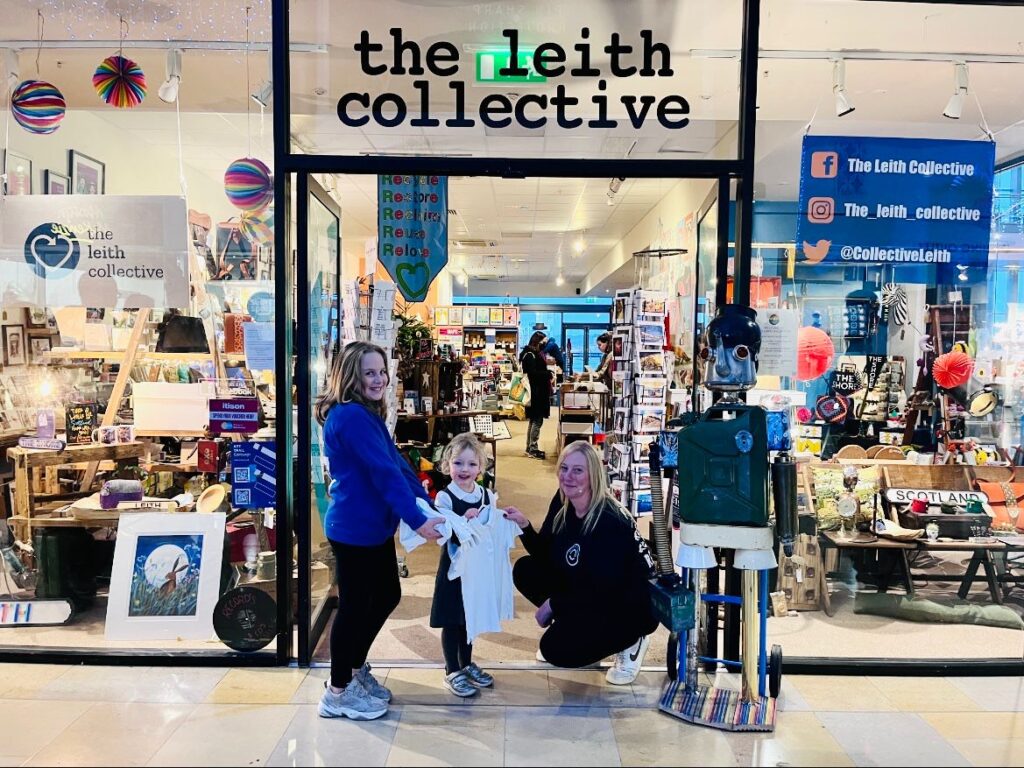 The Leith Collective School Uniform Exchange - a mum and two children stand outside the store, holding up school uniforms.