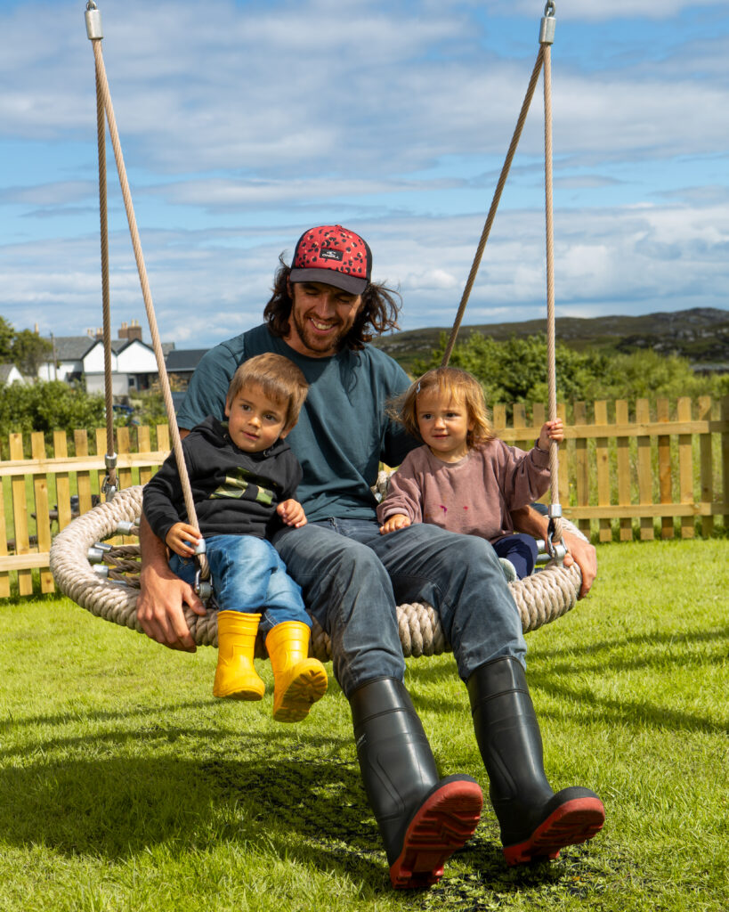 A dad and two children smile as they use a brand new basket swing in a green filled playpark.