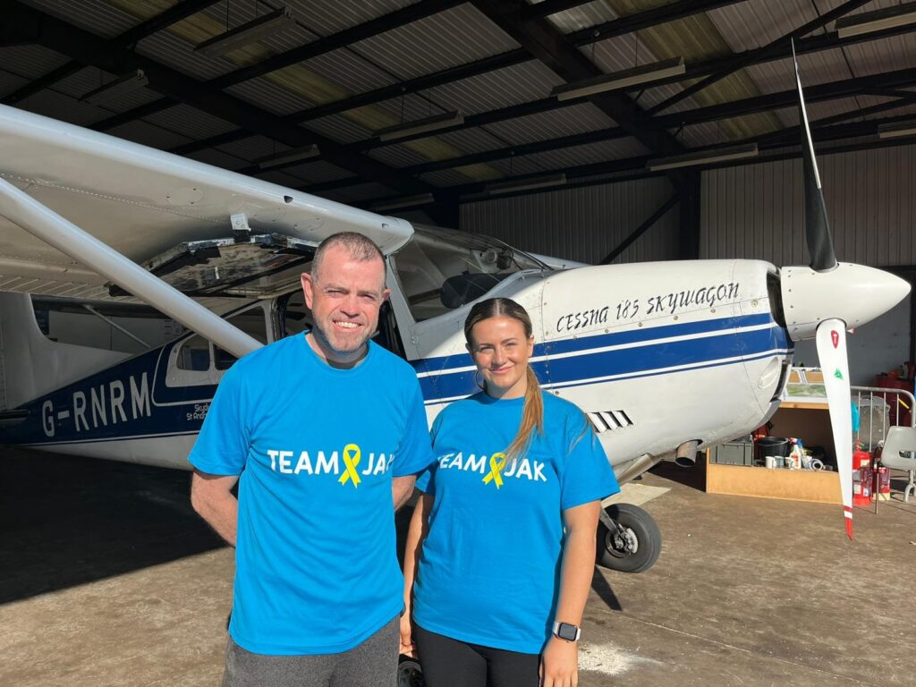 Lawyers Billy Smith and Chloe Herd stand in an aircraft hanger infront of a white aeroplane, ahead of their charity skydive