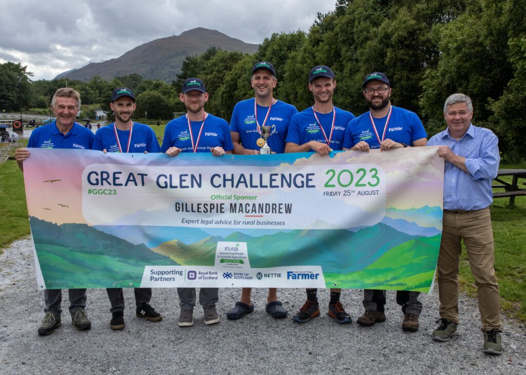 A team of athletes smile holding a long banner infront of their legs. The banner reads 'Great Glen Challenge 2023'.