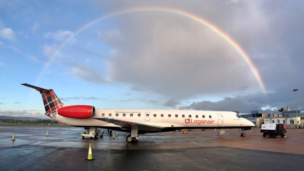 An aeroplane sits on the tarmac of an airport underneath a cloudy sky and a rainbow.