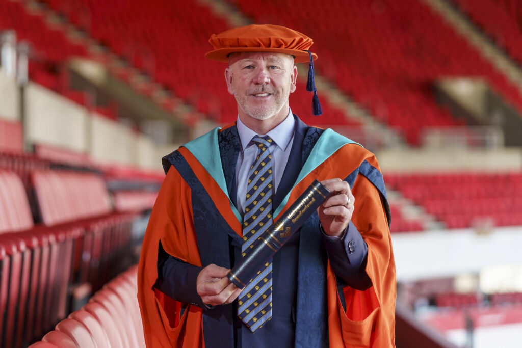 Kevin Ball in a graduate cap and gown, holding his degree scroll. Stadium of Light in the background