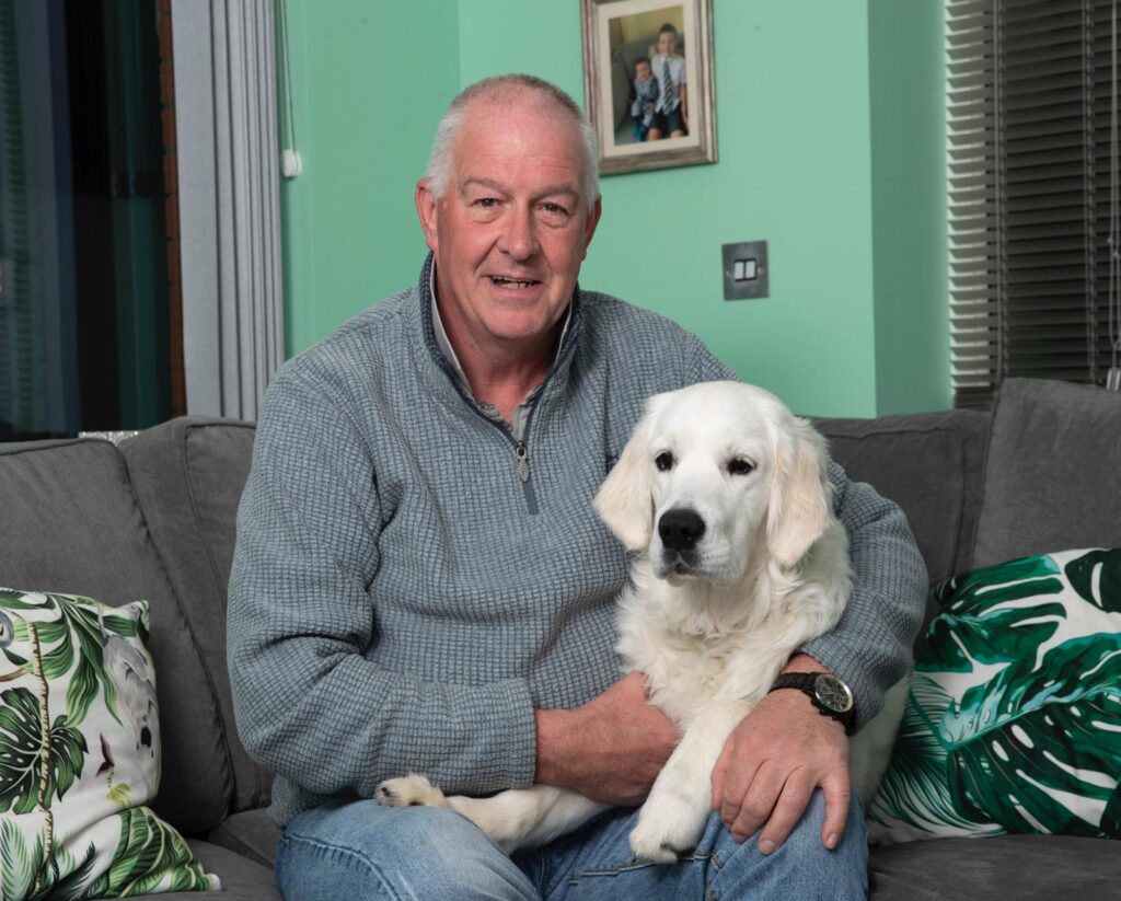 Rab Thomson smiles as he cuddles with white dog on couch.
