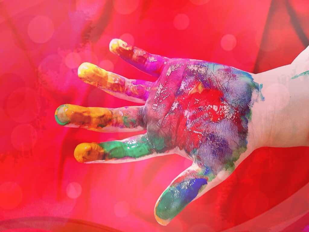 A hand covered in diffferent coloured paint in front of a red background.
