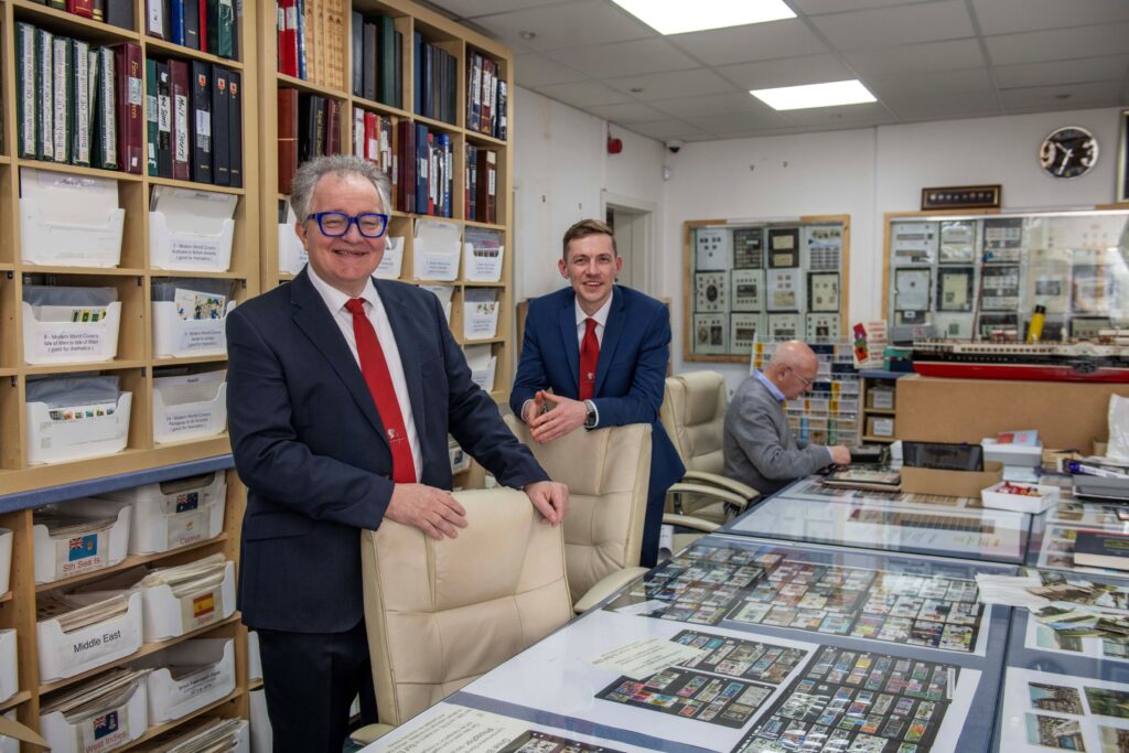 Glasgow Stamp Shop owner Gordon Carroll and son Austin Carroll smile for photo inside shop with customer in the back looking over stamps.