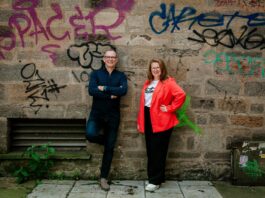 Kirsten Paul and Ray Bugg stand, smiling against a wall with graffiti. Image supplied with release by brass neck